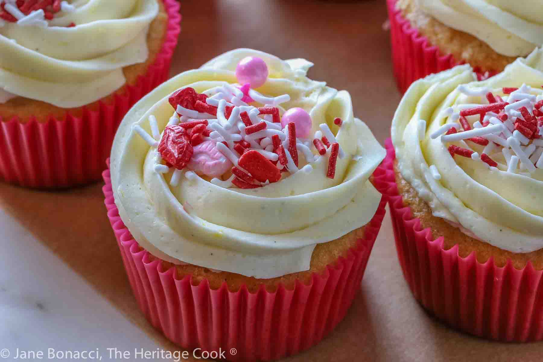 Multiple vanilla cupcakes in red paper liners, filled with whipped white chocolate ganache, topped with lemon kissed buttercream frosting and sprinkled with festive sprinkles © 2024 Jane Bonacci, The Heritage Cook. 