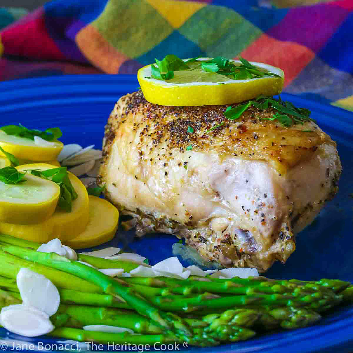 Large baked lemon chicken breast with a slice of lemon on top and green asparagus next to yellow squash on a blue plate © 2024 Jane Bonacci, The Heritage Cook.