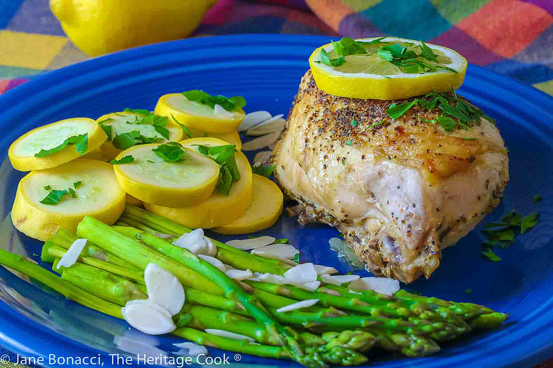Large baked lemon chicken breast with a slice of lemon on top and green asparagus next to yellow squash on a blue plate © 2024 Jane Bonacci, The Heritage Cook. 