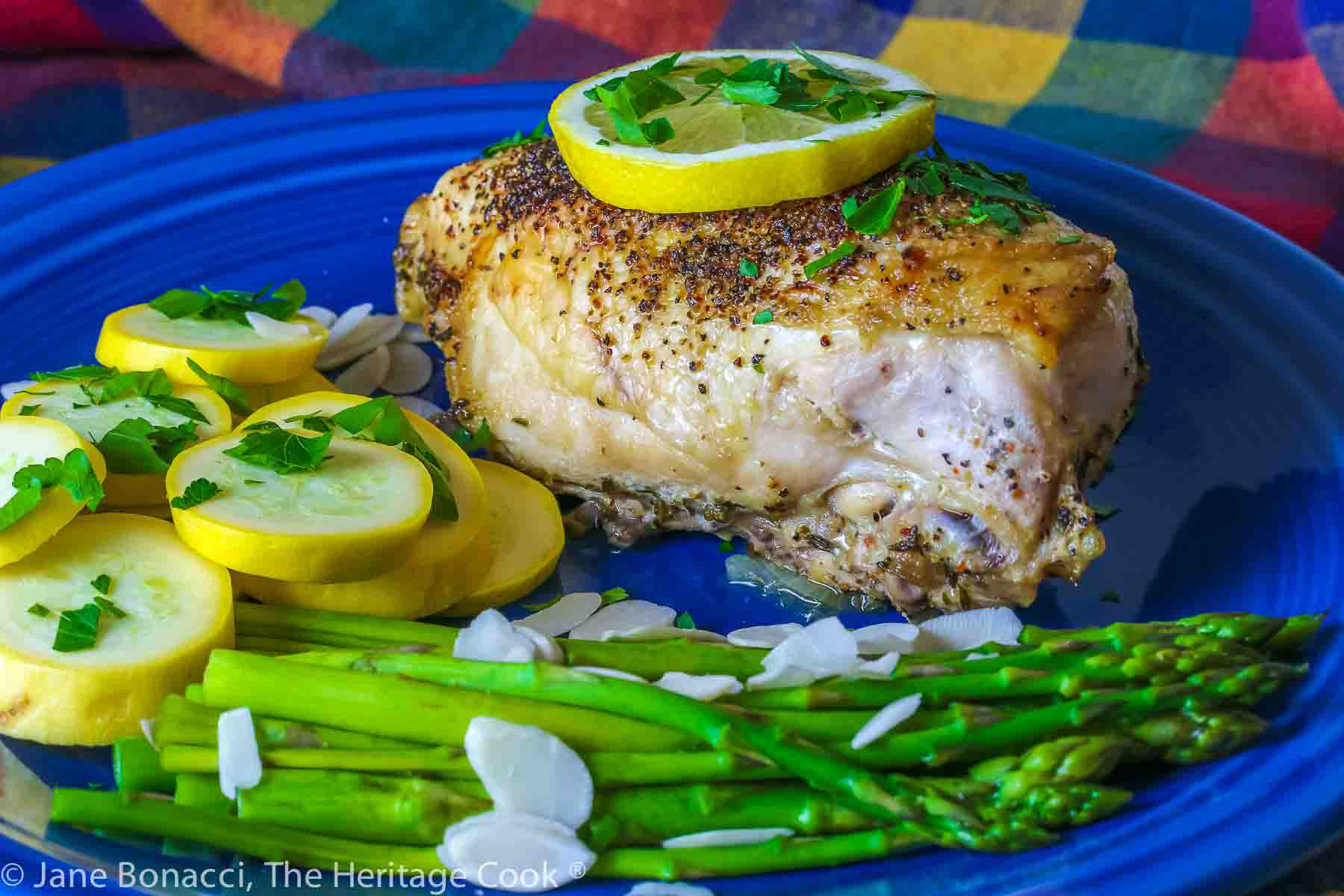 Large baked lemon chicken breast with a slice of lemon on top and green asparagus next to yellow squash on a blue plate © 2024 Jane Bonacci, The Heritage Cook. 