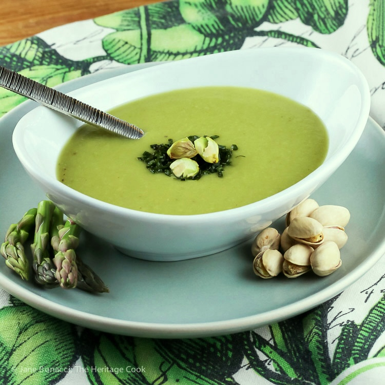 Green asparagus soup in a white oval bowl garnished with chives, chopped pistachios, and asparagus tips sitting on a green and white floral cloth © 2024 Jane Bonacci, The Heritage Cook.