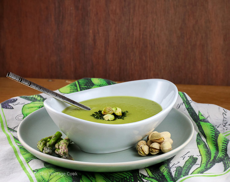 Green asparagus soup in a white oval bowl garnished with chives, chopped pistachios, and asparagus tips sitting on a green and white floral cloth © 2024 Jane Bonacci, The Heritage Cook. 