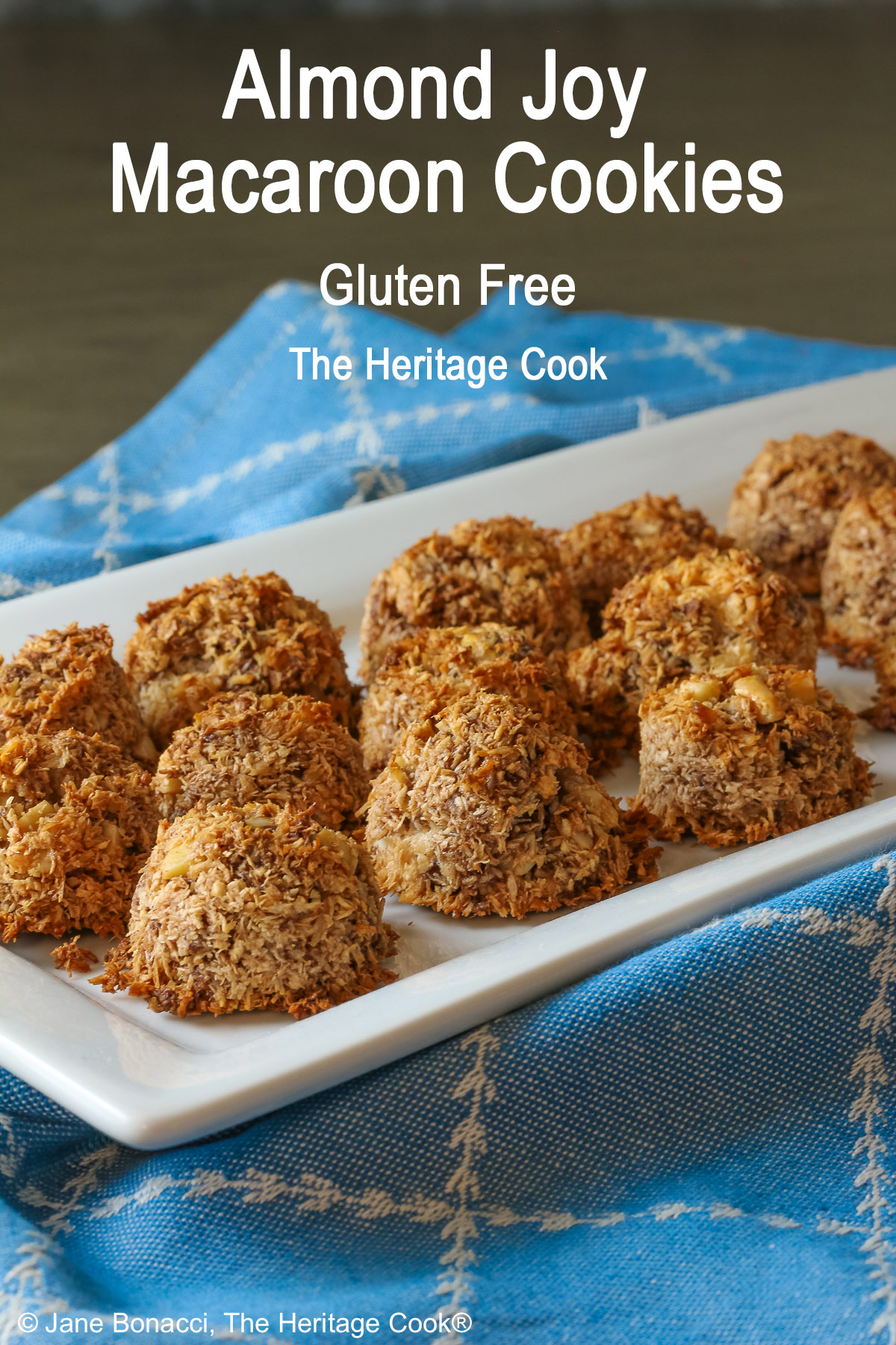 A white rectangular platter piled with coconut and chocolate chip Almond Joy Macaroon Cookies sitting on a blue cloth. They are gluten-free and dairy-free © 2024 Jane Bonacci, The Heritage Cook. 