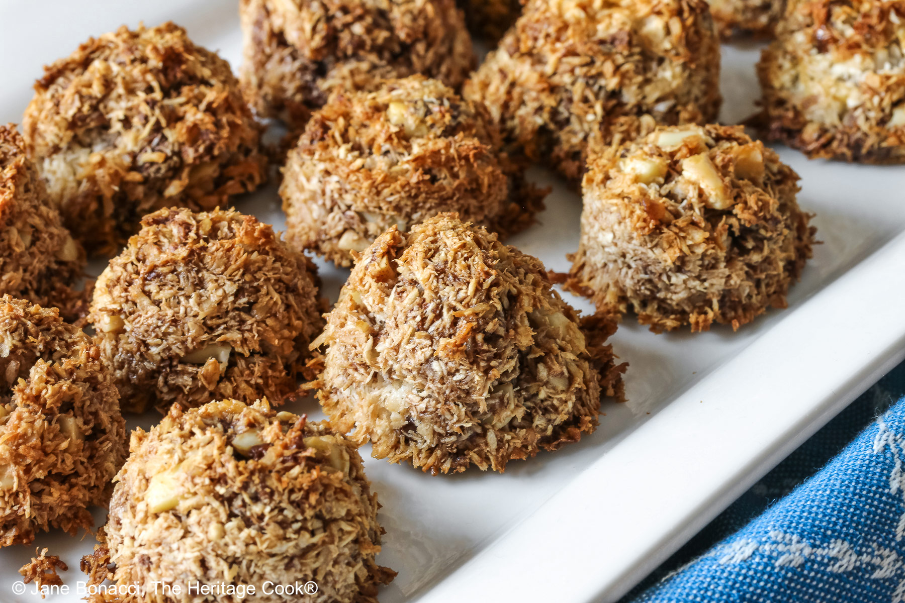 A white rectangular platter piled with coconut and chocolate chip Almond Joy Macaroon Cookies sitting on a blue cloth. They are gluten-free and dairy-free © 2024 Jane Bonacci, The Heritage Cook. 
