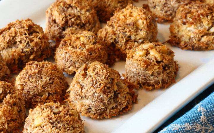 A white rectangular platter piled with coconut and chocolate chip Almond Joy Macaroon Cookies sitting on a blue cloth. They are gluten-free and dairy-free © 2024 Jane Bonacci, The Heritage Cook.