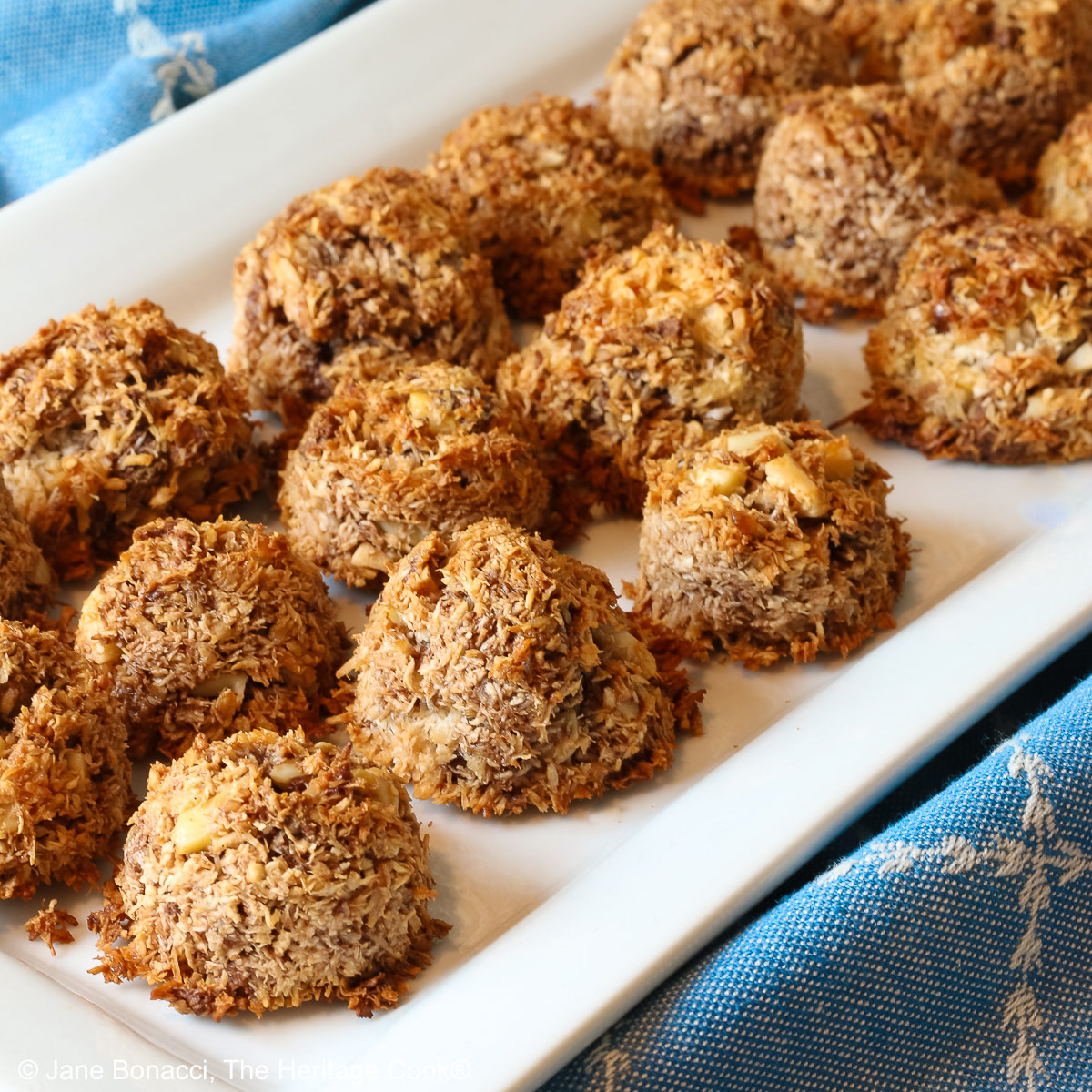 A white rectangular platter piled with coconut and chocolate chip Almond Joy Macaroon Cookies sitting on a blue cloth. They are gluten-free and dairy-free © 2024 Jane Bonacci, The Heritage Cook.