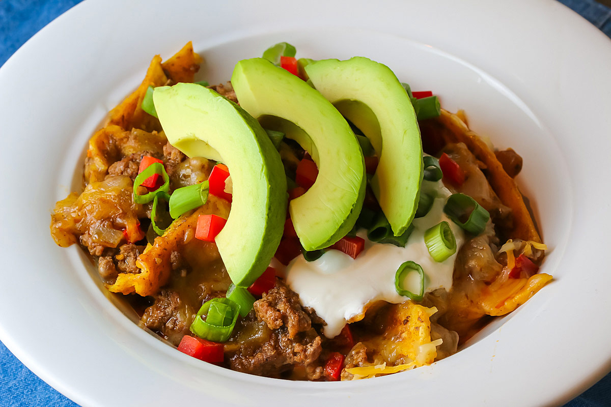 Cheesy Beef Taco One Pan Meal scooped into a white soup bowl and topped with green onions, red bell peppers, sour cream, and avocado slices sitting on a blue cloth © 2024 Jane Bonacci, The Heritage Cook.