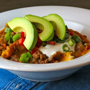 Cheesy Beef Taco One Pan Meal scooped into a white soup bowl and topped with green onions, red bell peppers, sour cream, and avocado slices sitting on a blue cloth © 2024 Jane Bonacci, The Heritage Cook.