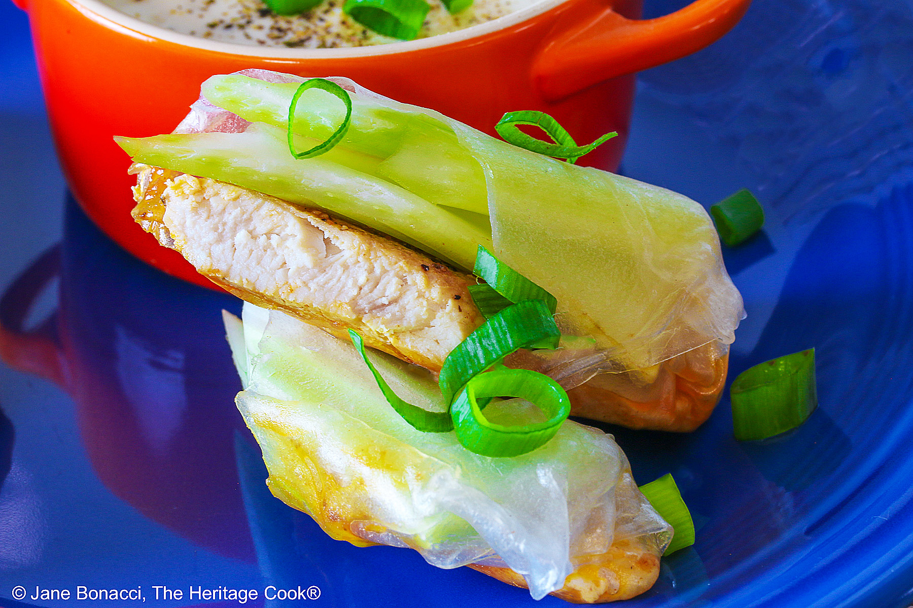 Buffalo Rice Paper Wraps with boneless chicken strips and celery sticks cut diagonally in half next to a bowl of blue cheese dip, both sprinkled with green onions, on a red and white checked cloth © 2024 Jane Bonacci, The Heritage Cook.
