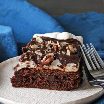 Mississippi Mud Brownies slice on a white plate topped with chocolate ganache and chopped pecans, with pecan halves and forks alongside, sitting on a blue cloth, some with an antique pitcher to the right of the plate © 2024 Jane Bonacci, The Heritage Cook.