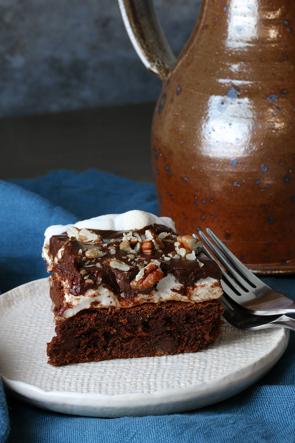 Mississippi Mud Brownies slice on a white plate topped with chocolate ganache and chopped pecans, with pecan halves and forks alongside, sitting on a blue cloth, some with an antique pitcher to the right of the plate © 2024 Jane Bonacci, The Heritage Cook. 