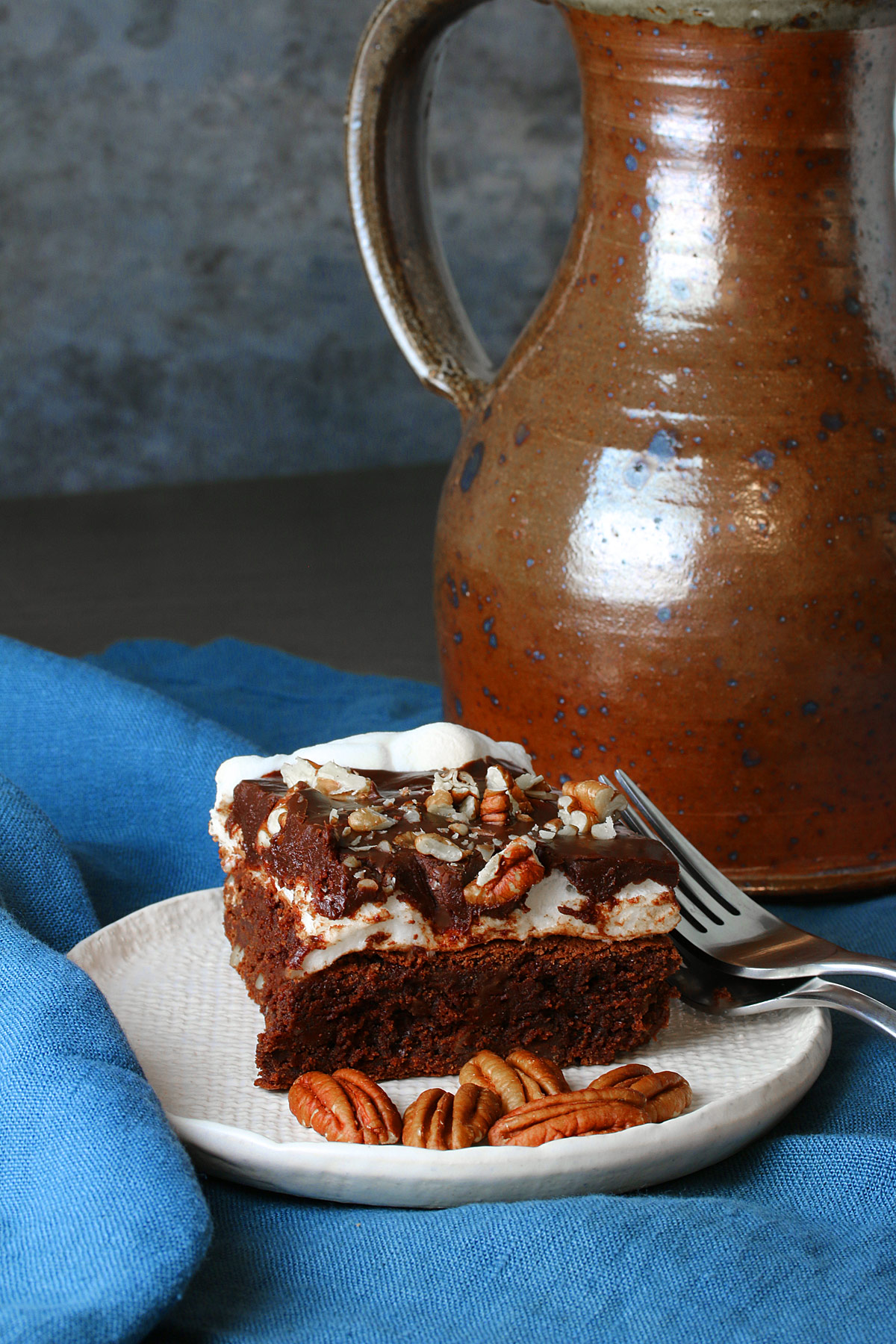 Mississippi Mud Brownies slice on a white plate topped with chocolate ganache and chopped pecans, with pecan halves and forks alongside, sitting on a blue cloth, some with an antique pitcher to the right of the plate © 2024 Jane Bonacci, The Heritage Cook. 