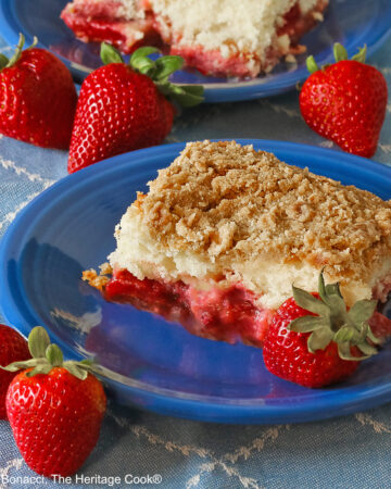 Two slices of Strawberry White Chocolate Crumb Cake on two blue plates sitting on a pale blue cloth with whole strawberries around the plates © 2024 Jane Bonacci, The Heritage Cook.