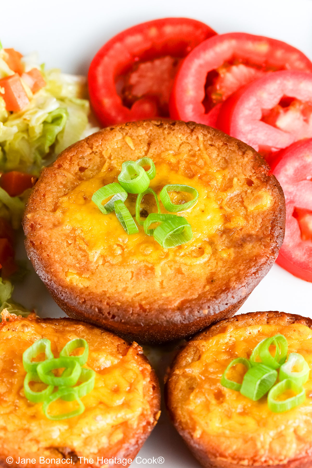 Golden brown Cheesy Sloppy Joe Tamale Cups with sliced green onions on top on a white plate with colorful coleslaw and bright red tomato slices sitting on a red and white cloth © 2026 Jane Bonacci, The Heritage Cook.
