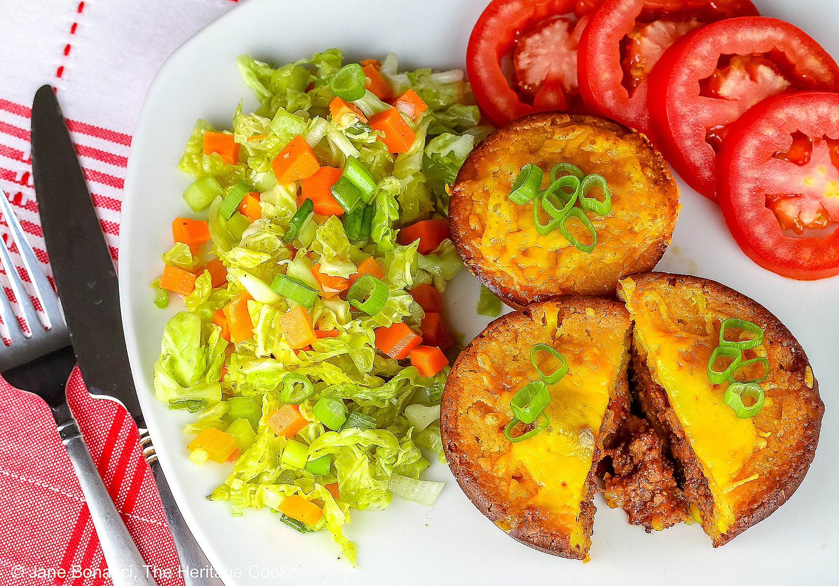Golden brown Cheesy Sloppy Joe Tamale Cups with sliced green onions on top on a white plate with colorful coleslaw and bright red tomato slices sitting on a red and white cloth © 2026 Jane Bonacci, The Heritage Cook.