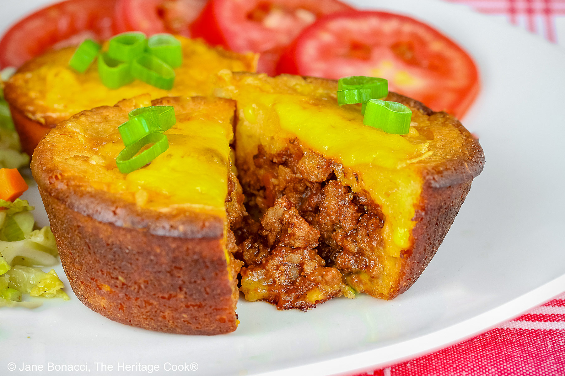 Golden brown Cheesy Sloppy Joe Tamale Cups with sliced green onions on top on a white plate with colorful coleslaw and bright red tomato slices sitting on a red and white cloth © 2026 Jane Bonacci, The Heritage Cook.