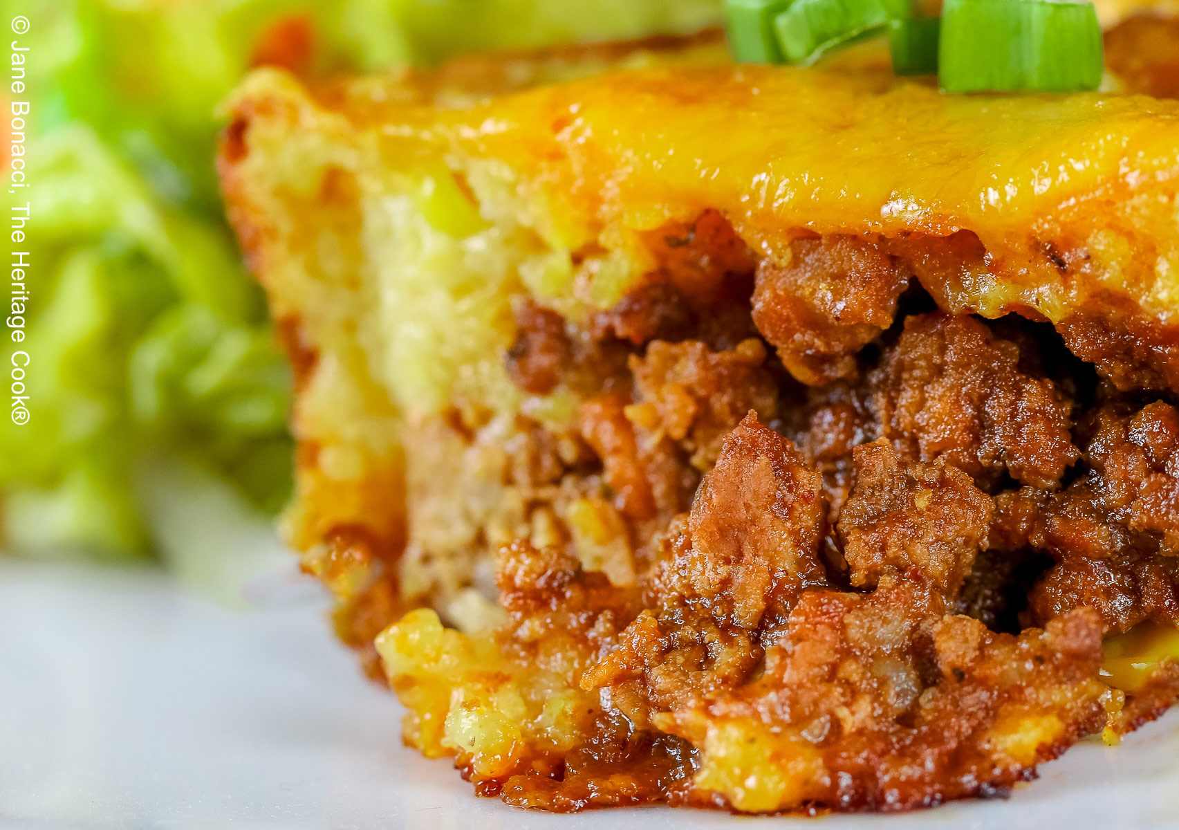 Golden brown Cheesy Sloppy Joe Tamale Cups with sliced green onions on top on a white plate with colorful coleslaw and bright red tomato slices sitting on a red and white cloth © 2026 Jane Bonacci, The Heritage Cook.