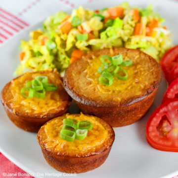 Golden brown Cheesy Sloppy Joe Tamale Cups with sliced green onions on top on a white plate with colorful coleslaw and bright red tomato slices sitting on a red and white cloth © 2026 Jane Bonacci, The Heritage Cook.