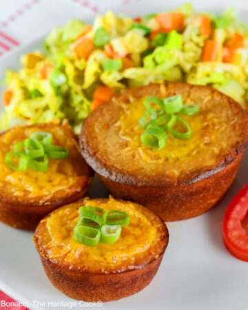 Golden brown Cheesy Sloppy Joe Tamale Cups with sliced green onions on top on a white plate with colorful coleslaw and bright red tomato slices sitting on a red and white cloth © 2026 Jane Bonacci, The Heritage Cook.