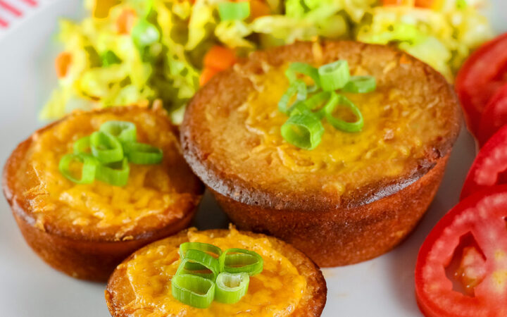 Golden brown Cheesy Sloppy Joe Tamale Cups with sliced green onions on top on a white plate with colorful coleslaw and bright red tomato slices sitting on a red and white cloth © 2026 Jane Bonacci, The Heritage Cook.