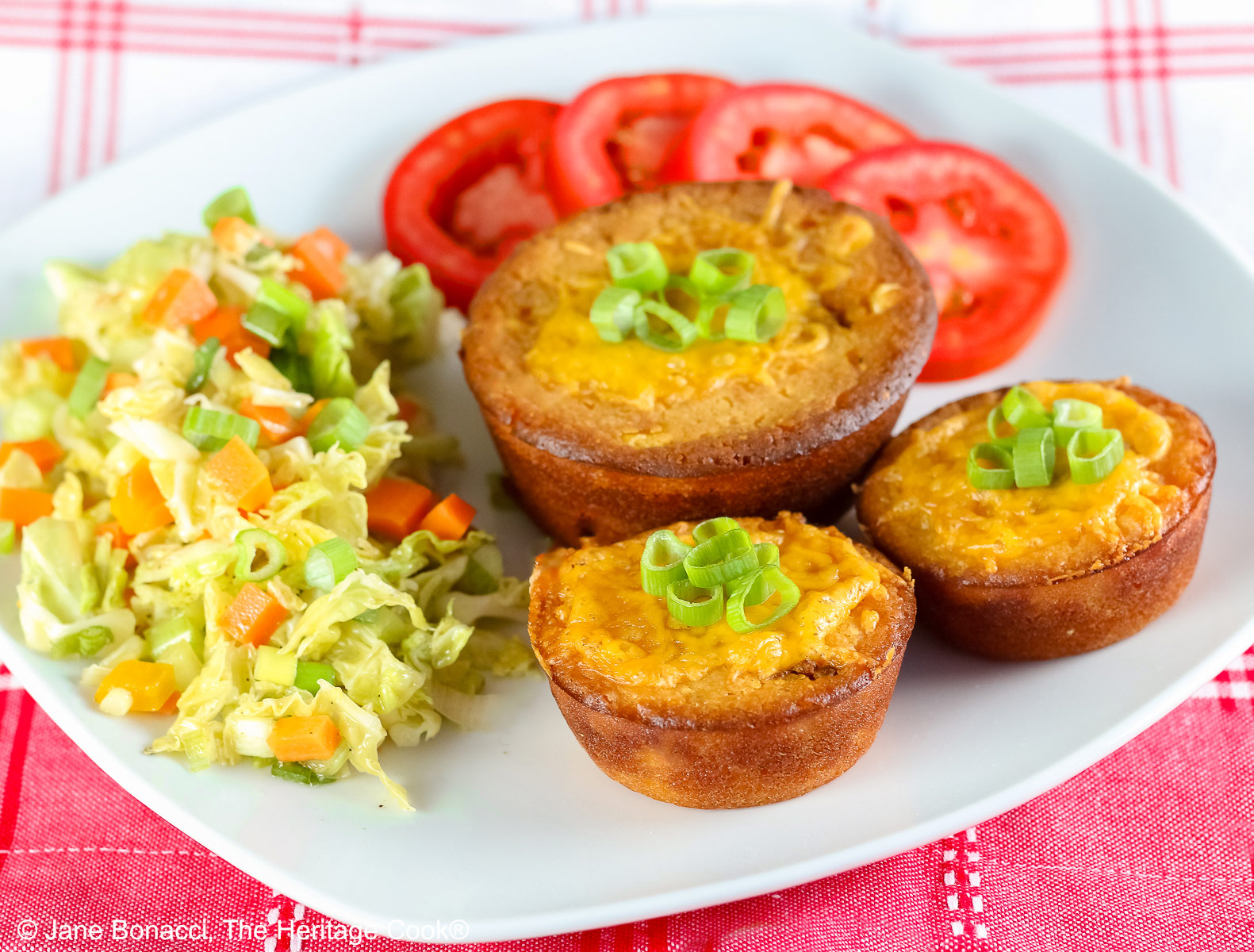Golden brown Cheesy Sloppy Joe Tamale Cups with sliced green onions on top on a white plate with colorful coleslaw and bright red tomato slices sitting on a red and white cloth © 2026 Jane Bonacci, The Heritage Cook.