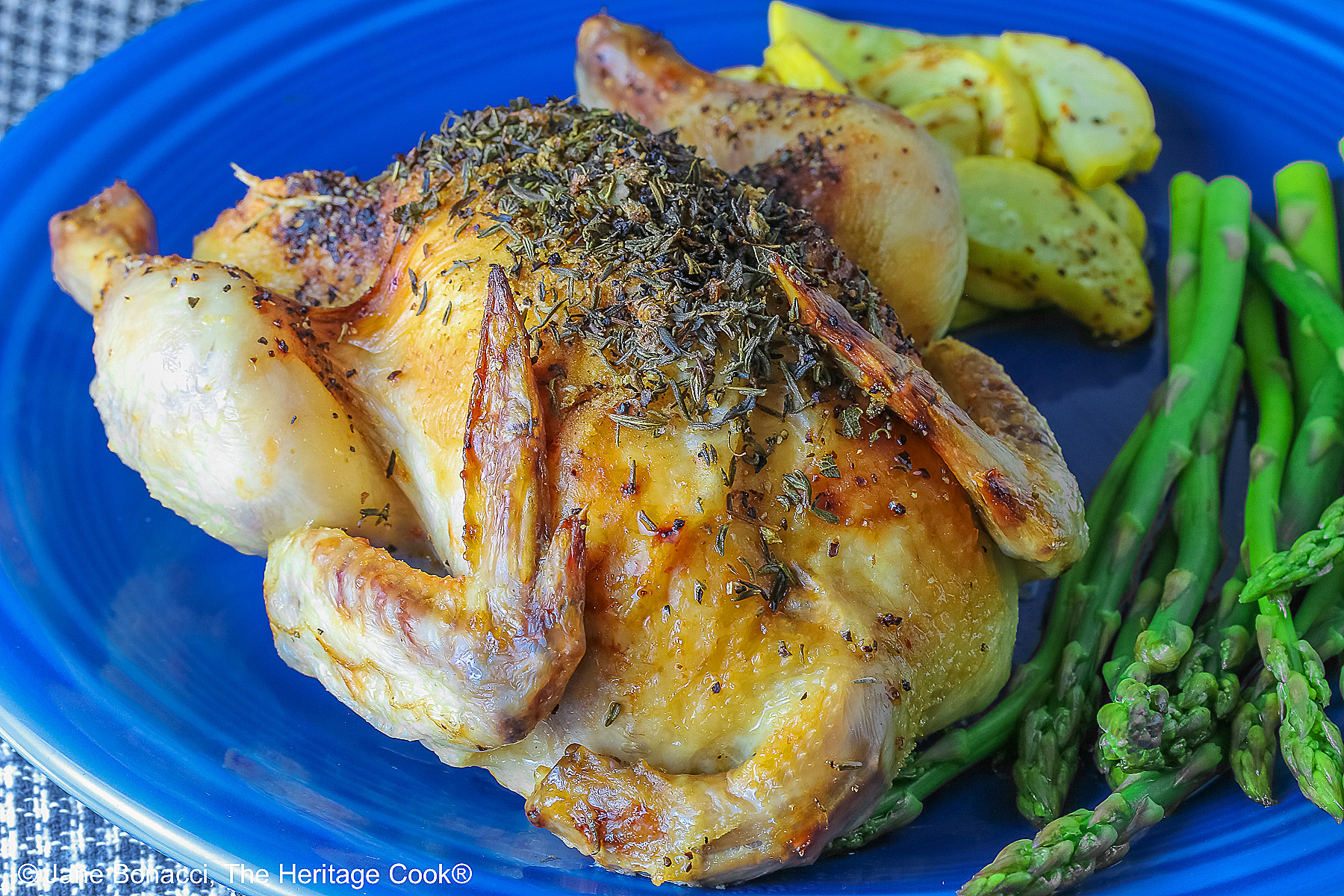 A Roasted Game Hen with Herb Butter for Valentine's Day on a deep blue plate with sauteed yellow squash and steamed asparagus, sitting on a checked cloth © 2025 Jane Bonacci, The Heritage Cook. 