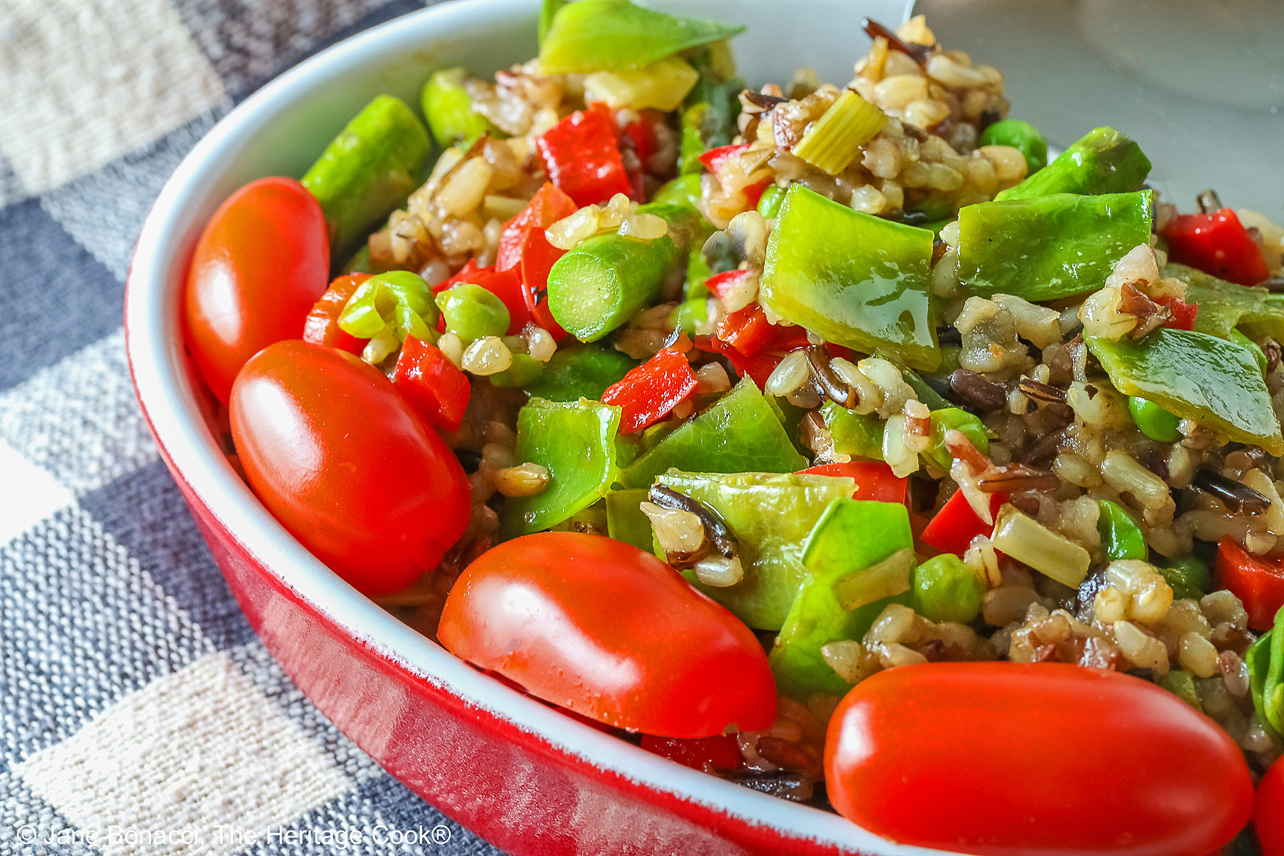 Brown & Wild Rice Pilaf piled into a red au gratin dish stuffed with lots of green vegetables and ringed with red cherry tomatoes, with a spoon tucked into the back of the dish, sitting on a grey and white checked cloth © 2025 Jane Bonacci, The Heritage Cook. 