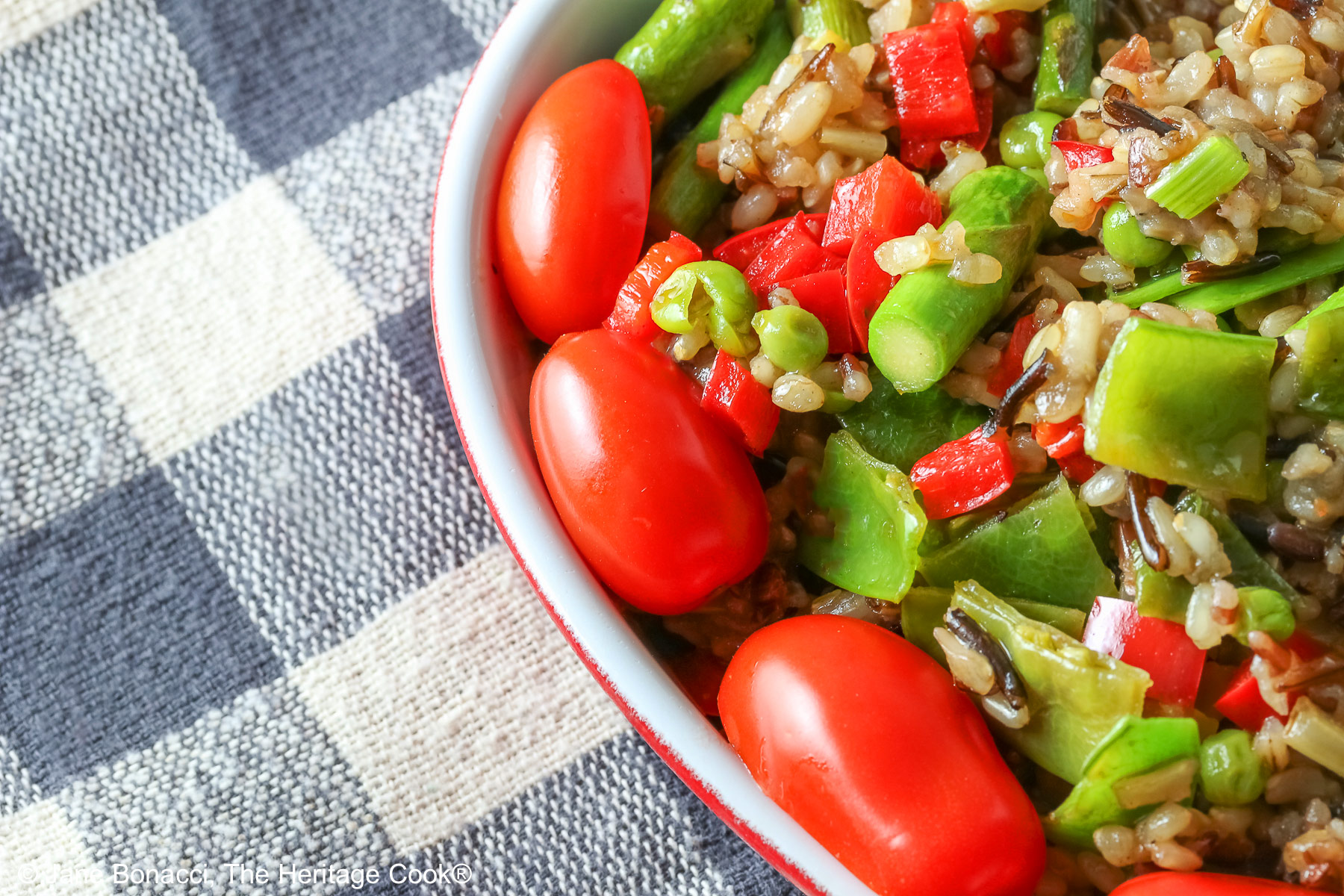Brown & Wild Rice Pilaf piled into a red au gratin dish stuffed with lots of green vegetables and ringed with red cherry tomatoes, with a spoon tucked into the back of the dish, sitting on a grey and white checked cloth © 2025 Jane Bonacci, The Heritage Cook. 