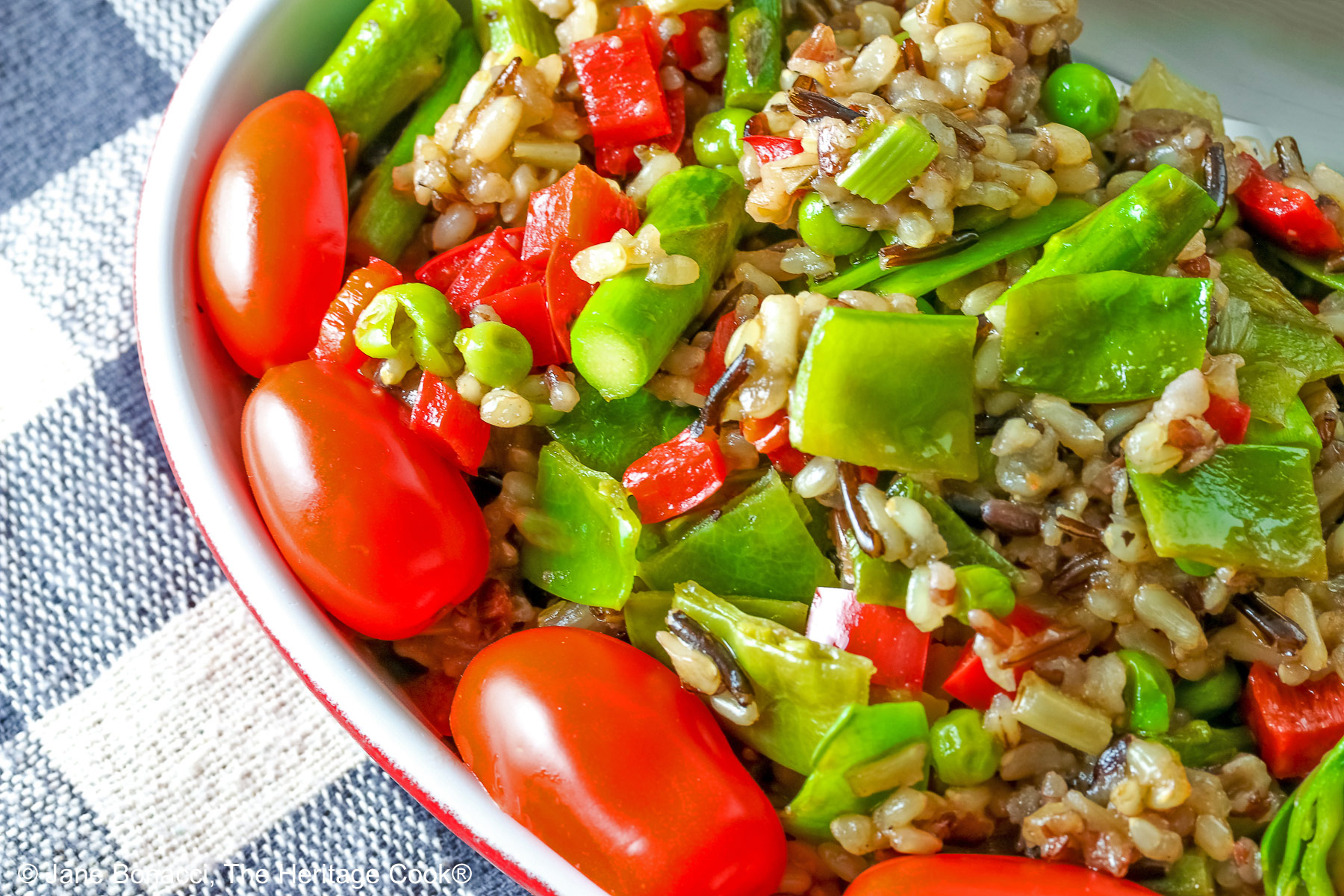 Brown & Wild Rice Pilaf piled into a red au gratin dish stuffed with lots of green vegetables and ringed with red cherry tomatoes, with a spoon tucked into the back of the dish, sitting on a grey and white checked cloth © 2025 Jane Bonacci, The Heritage Cook. 