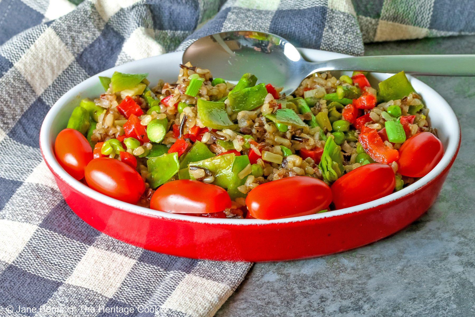 Brown & Wild Rice Pilaf piled into a red au gratin dish stuffed with lots of green vegetables and ringed with red cherry tomatoes, with a spoon tucked into the back of the dish, sitting on a grey and white checked cloth © 2025 Jane Bonacci, The Heritage Cook. 