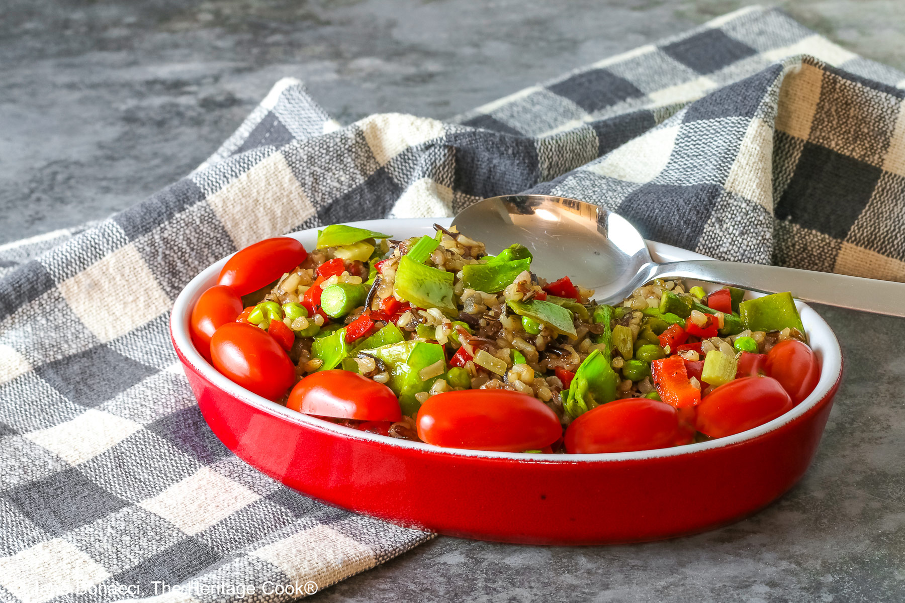 Brown & Wild Rice Pilaf piled into a red au gratin dish stuffed with lots of green vegetables and ringed with red cherry tomatoes, with a spoon tucked into the back of the dish, sitting on a grey and white checked cloth © 2025 Jane Bonacci, The Heritage Cook. 