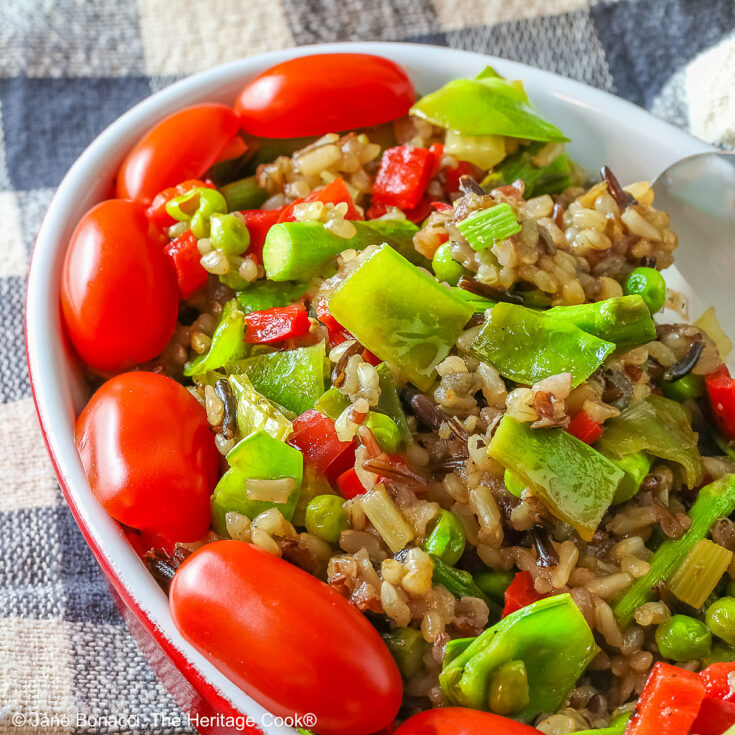 Brown & Wild Rice Pilaf piled into a red au gratin dish stuffed with lots of green vegetables and ringed with red cherry tomatoes, with a spoon tucked into the back of the dish, sitting on a grey and white checked cloth © 2025 Jane Bonacci, The Heritage Cook.