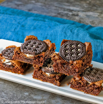 Line of 3 Decadent Oreo Brownies with two staggered and stacked on top showing the Oreo cookies baked into the top of the brownies on a white rectangular plate with a blue cloth behind © 2025 Jane Bonacci, The Heritage Cook.