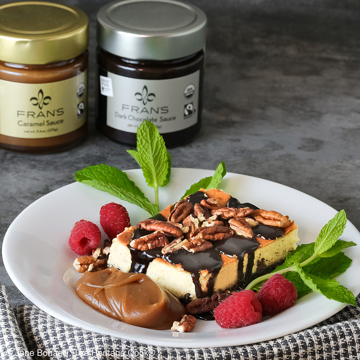 A slice of Turtle Cheesecake Bars on a white plate topped with chocolate sauce and chopped pecans with a dollop of caramel sauce on the plate surrounded by more nuts, raspberries, and mint sprigs © 2025 Jane Bonacci, The Heritage Cook.