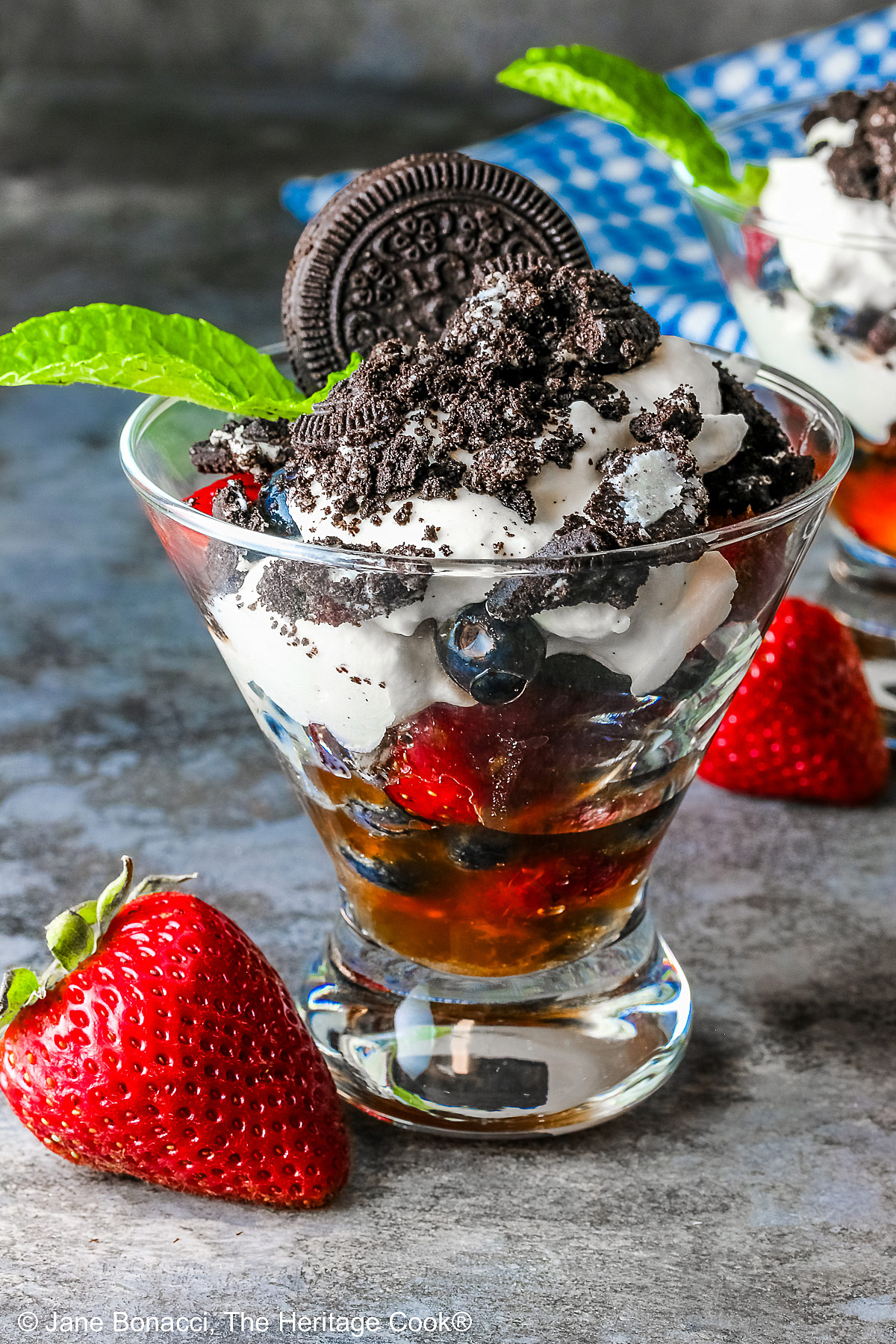 Fresh Berry Oreo Cream Parfaits (GF) in glass dishes to show the beautiful layers of fruit, whipped cream, and crushed cookies with a mint sprig and a whole cookie on top with a blue and white checked cloth in the background © 2025 Jane Bonacci, The Heritage Cook.
