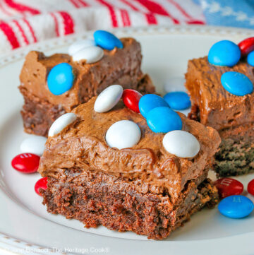 Three frosted fudgy brownies on a white plate topped with red, white, and blue M&M candies, some extra candies scattered on the plate and red, white, and blue cloths behind the plate © 2025 Jane Bonacci, The Heritage Cook.