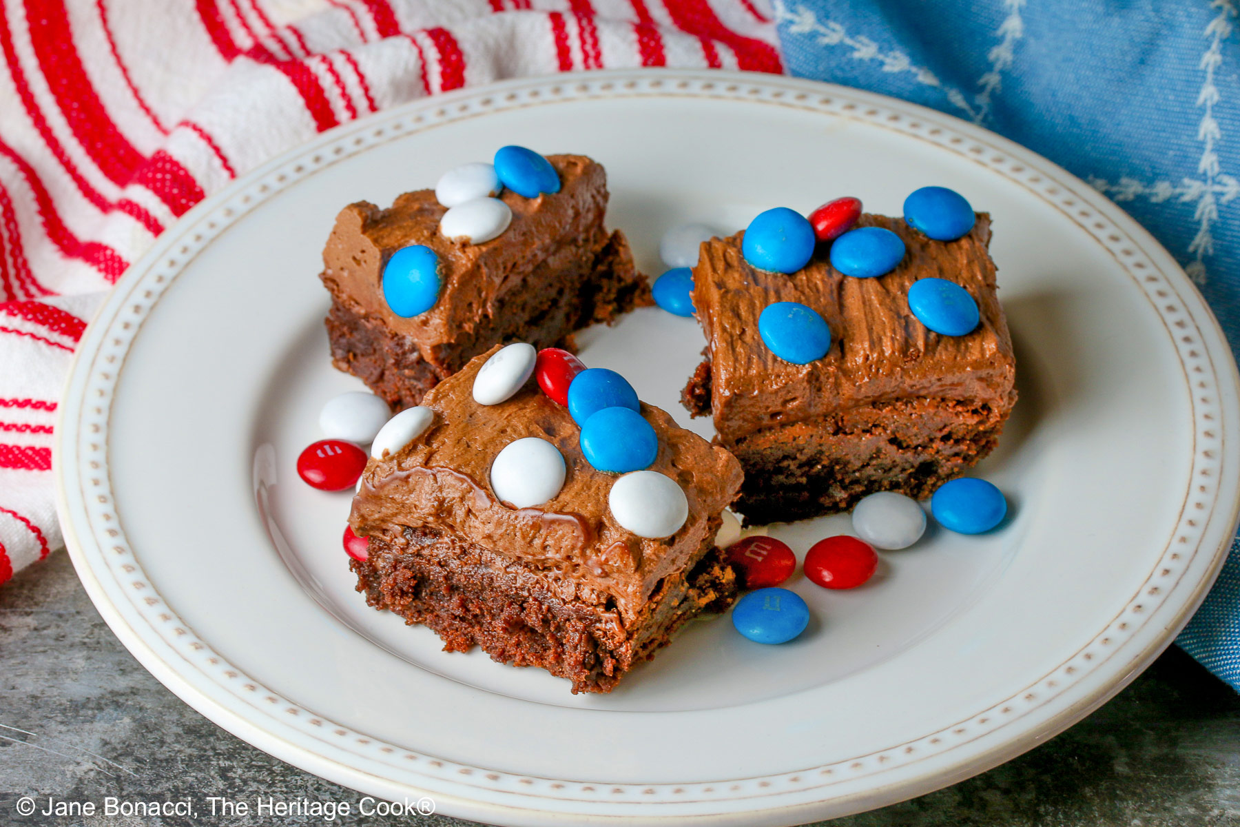Three frosted fudgy brownies on a white plate topped with red, white, and blue M&M candies, some extra candies scattered on the plate and red, white, and blue cloths behind the plate © 2025 Jane Bonacci, The Heritage Cook.