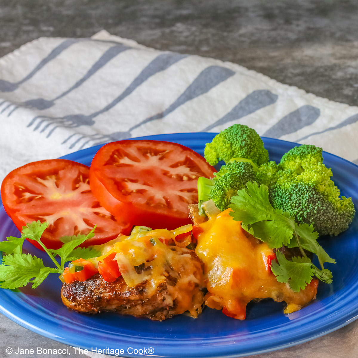 Tex Mex Chicken Supper served on a dark blue plate with two pieces of cheese covered chicken, sliced red tomatoes, bright green broccoli, and green cilantro leaves, with a cloth beneath and behind, some with a fork on the back right of the plate © 2025 Jane Bonacci, The Heritage Cook.
