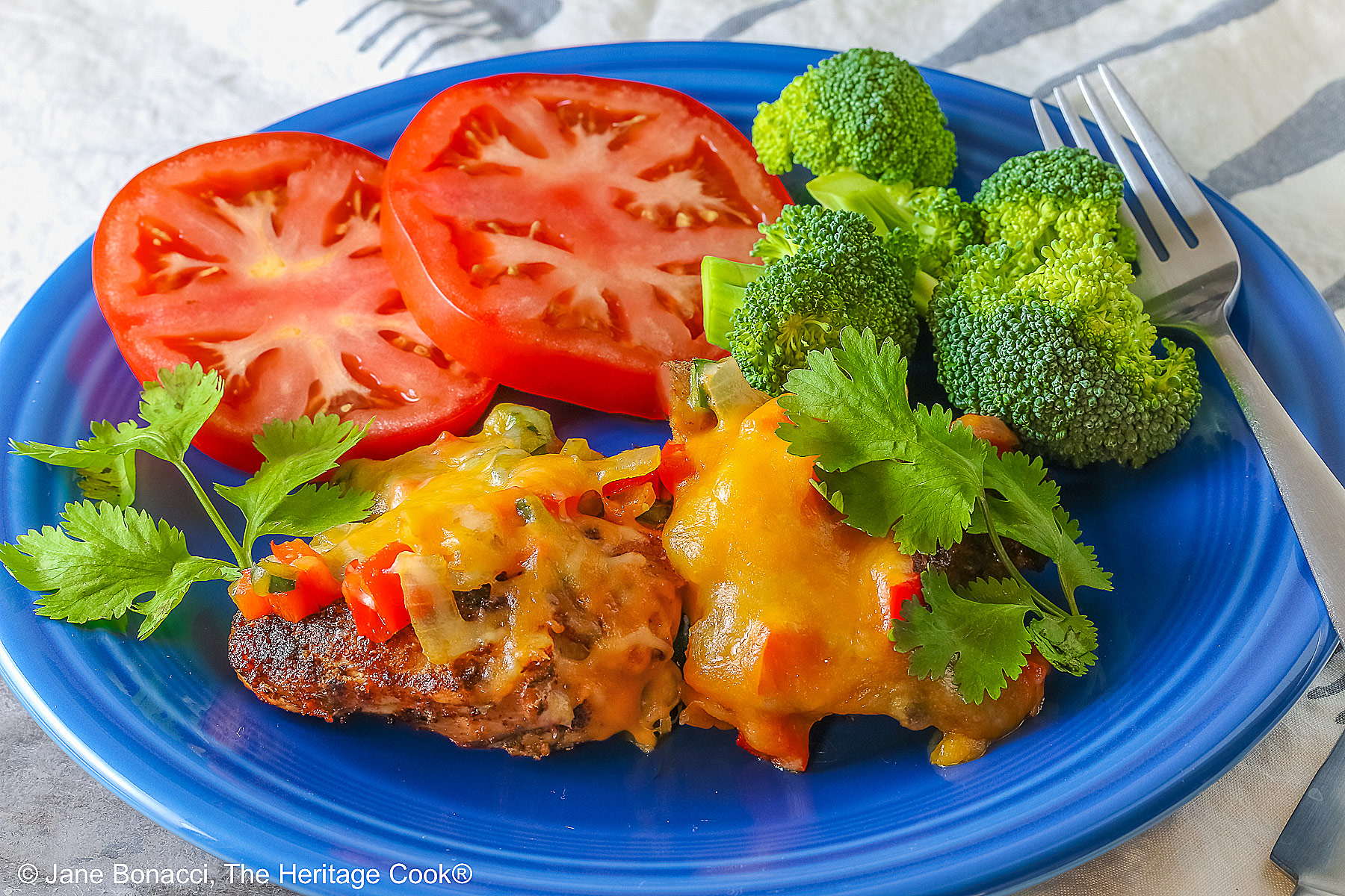 Tex Mex Chicken Supper served on a dark blue plate with two pieces of cheese covered chicken, sliced red tomatoes, bright green broccoli, and green cilantro leaves, with a cloth beneath and behind, some with a fork on the back right of the plate © 2025 Jane Bonacci, The Heritage Cook. 