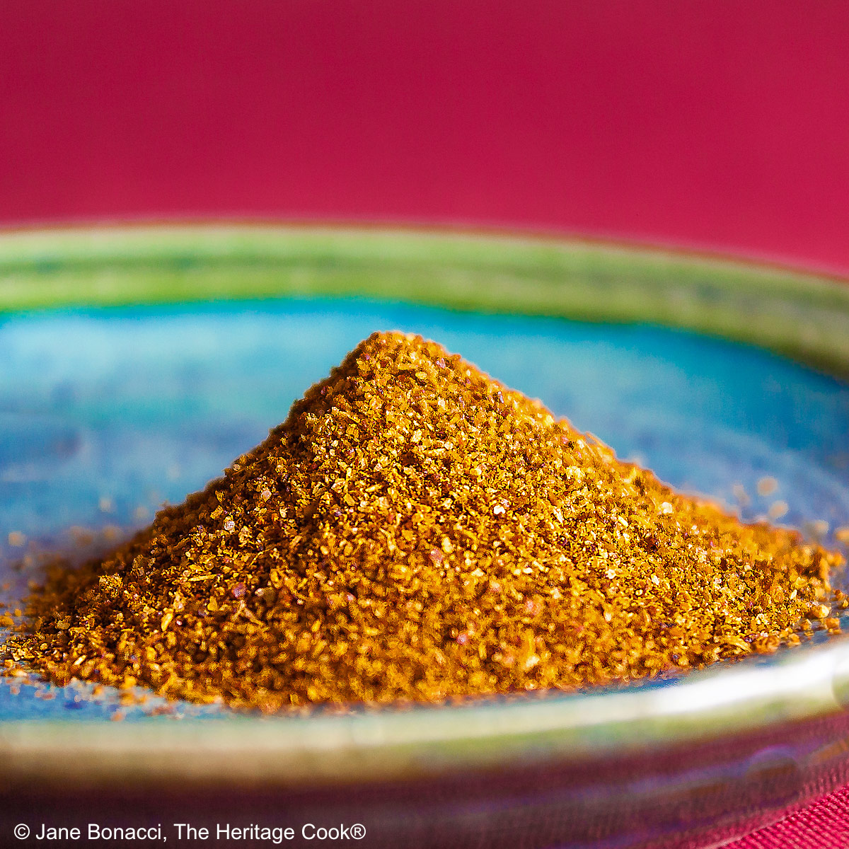 Redish-golden homemade BBQ dry rub piled on a small blue plate with a green rim, sitting on a dark red cloth © 2025 Jane Bonacci, The Heritage Cook.