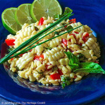 Bowl of lovely yellow Corn, Orzo, and Basil Salad with Lime Vinaigrette studded with red bell peppers and chopped basil in a dark blue bowl; topped with a sprig of basil, some chives, and 3 lime slices on a light blue cloth © 2025 Jane Bonacci, The Heritage Cook.