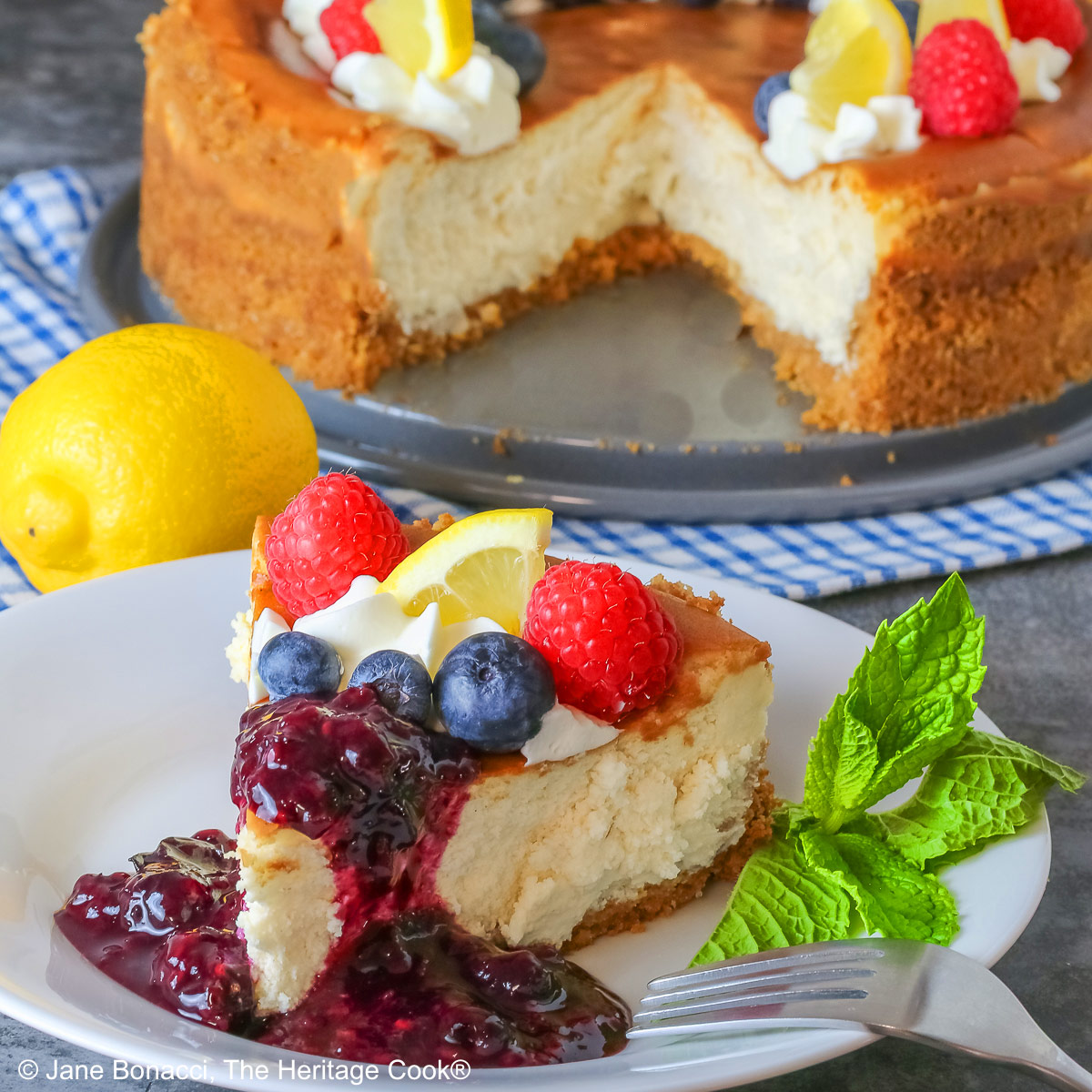Slice of Lemon Cheesecake with Fresh Berry Sauce on a white plate topped with raspberries, blueberries, piped whipped cream and a mini lemon wedge and the berry sauce poured over the tip of the slice, sitting on a blue and white checked cloth © 2025 Jane Bonacci, The Heritage Cook.