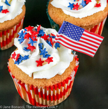 Vanilla cupcakes in flag-decorated paper liners, topped with a crown of white chocolate frosting sprinkled with red, white, and blue star candies with a miniature American flag flying on the top © 2025 Jane Bonacci, The Heritage Cook.
