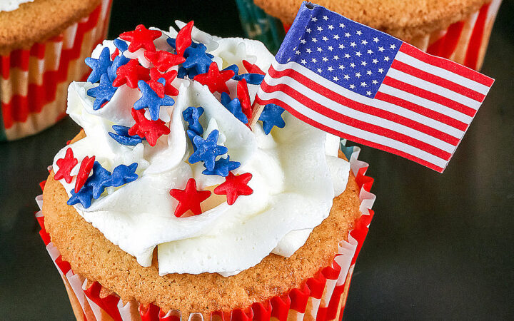 Macadamia Nut White Chocolate Cupcakes (GF) in American flag paper liners, topped with swirled white buttercream frosting, sprinkled with red and blue candy stars with a mini American flag inserted in each cupcake, sitting on a black background with a blue and white checked cloth © 2026 Jane Bonacci, The Heritage Cook.