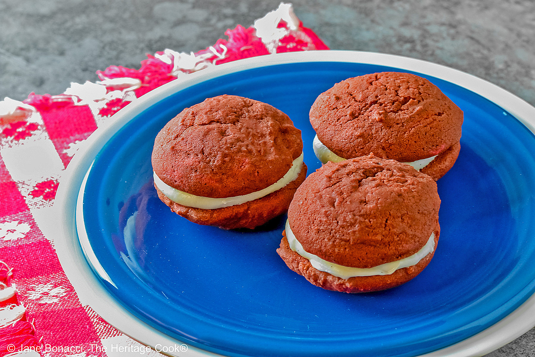 3 Mini Red Velvet Whoopie Pies clustered together in the center of a small dark blue plate on top of a larger white plate, sitting on a red and white checked cloth, perfect for the upcoming 4th of July parties © 2025 Jane Bonacci, The Heritage Cook.