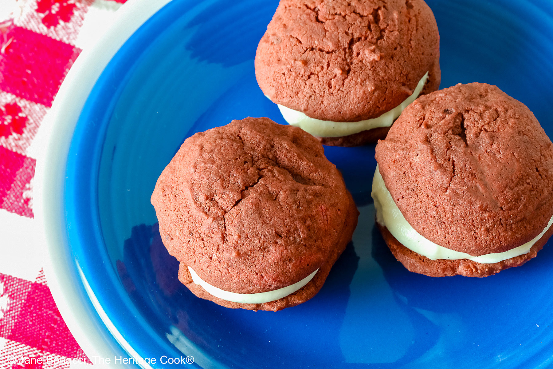 3 Mini Red Velvet Whoopie Pies clustered together in the center of a small dark blue plate on top of a larger white plate, sitting on a red and white checked cloth, perfect for the upcoming 4th of July parties © 2025 Jane Bonacci, The Heritage Cook.