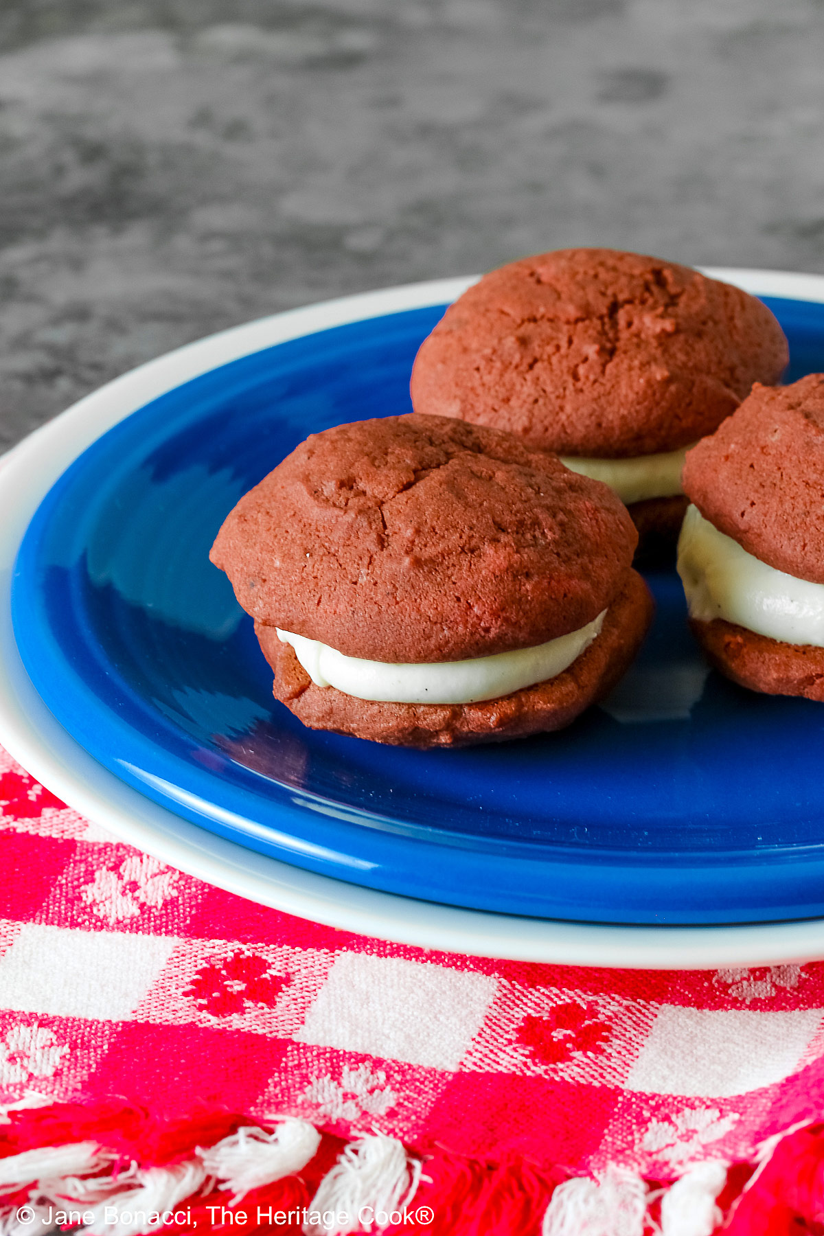 3 Mini Red Velvet Whoopie Pies clustered together in the center of a small dark blue plate on top of a larger white plate, sitting on a red and white checked cloth, perfect for the upcoming 4th of July parties © 2025 Jane Bonacci, The Heritage Cook.