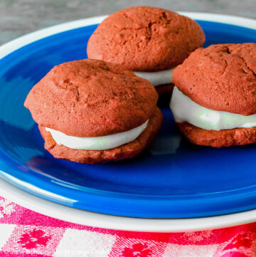 3 Mini Red Velvet Whoopie Pies clustered together in the center of a small dark blue plate on top of a larger white plate, sitting on a red and white checked cloth, perfect for the upcoming 4th of July parties © 2025 Jane Bonacci, The Heritage Cook.