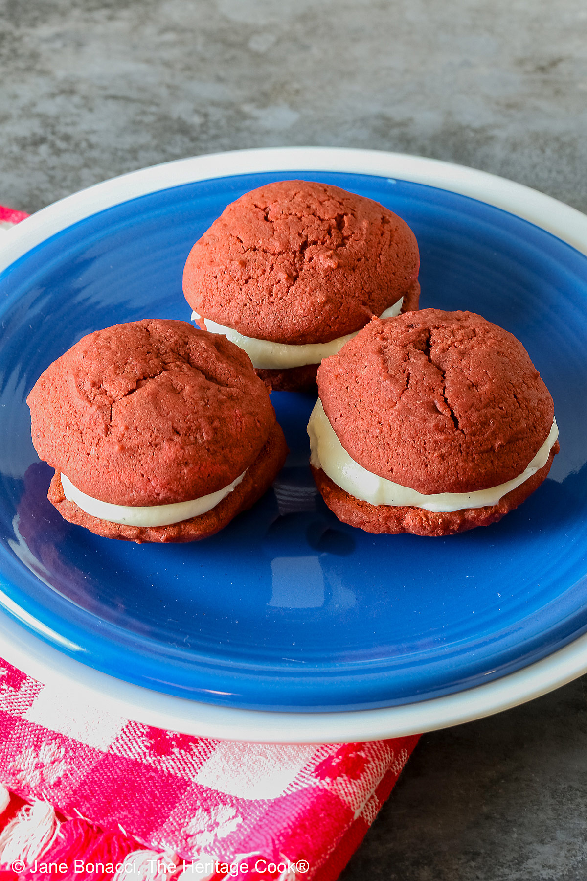 3 Mini Red Velvet Whoopie Pies clustered together in the center of a small dark blue plate on top of a larger white plate, sitting on a red and white checked cloth, perfect for the upcoming 4th of July parties © 2025 Jane Bonacci, The Heritage Cook.