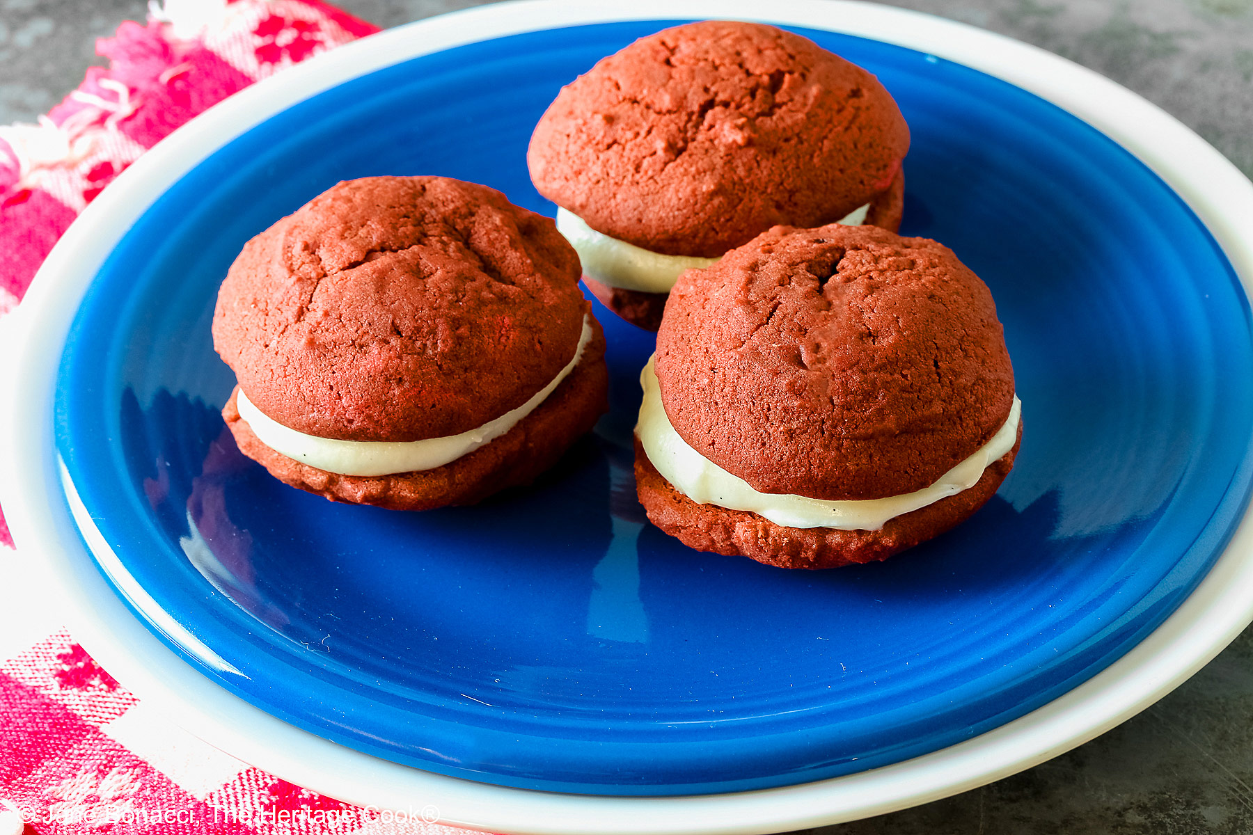 3 Mini Red Velvet Whoopie Pies clustered together in the center of a small dark blue plate on top of a larger white plate, sitting on a red and white checked cloth, perfect for the upcoming 4th of July parties © 2025 Jane Bonacci, The Heritage Cook.